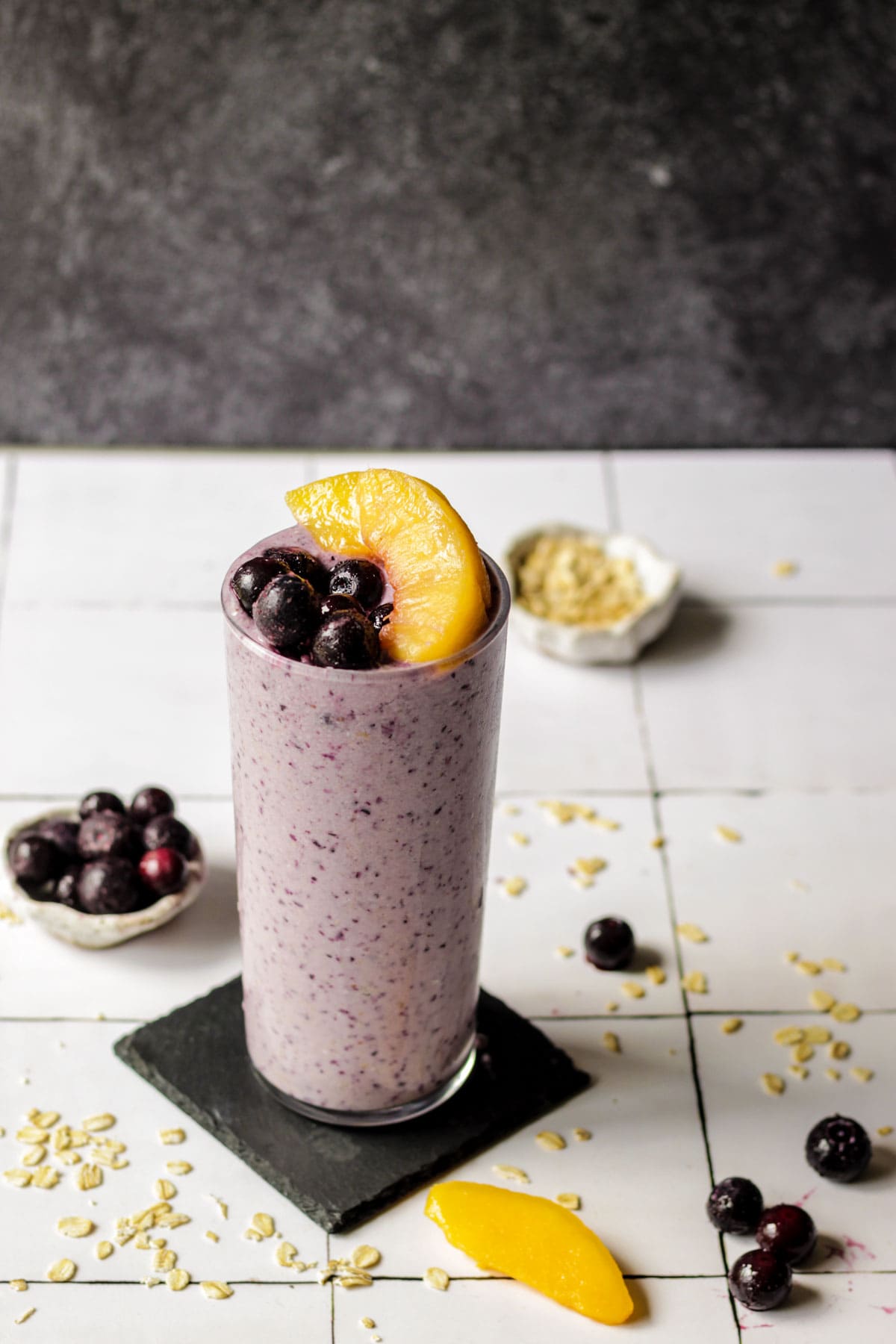 A glass of blueberry peach oatmeal smoothie with bowls of blueberries and peaches