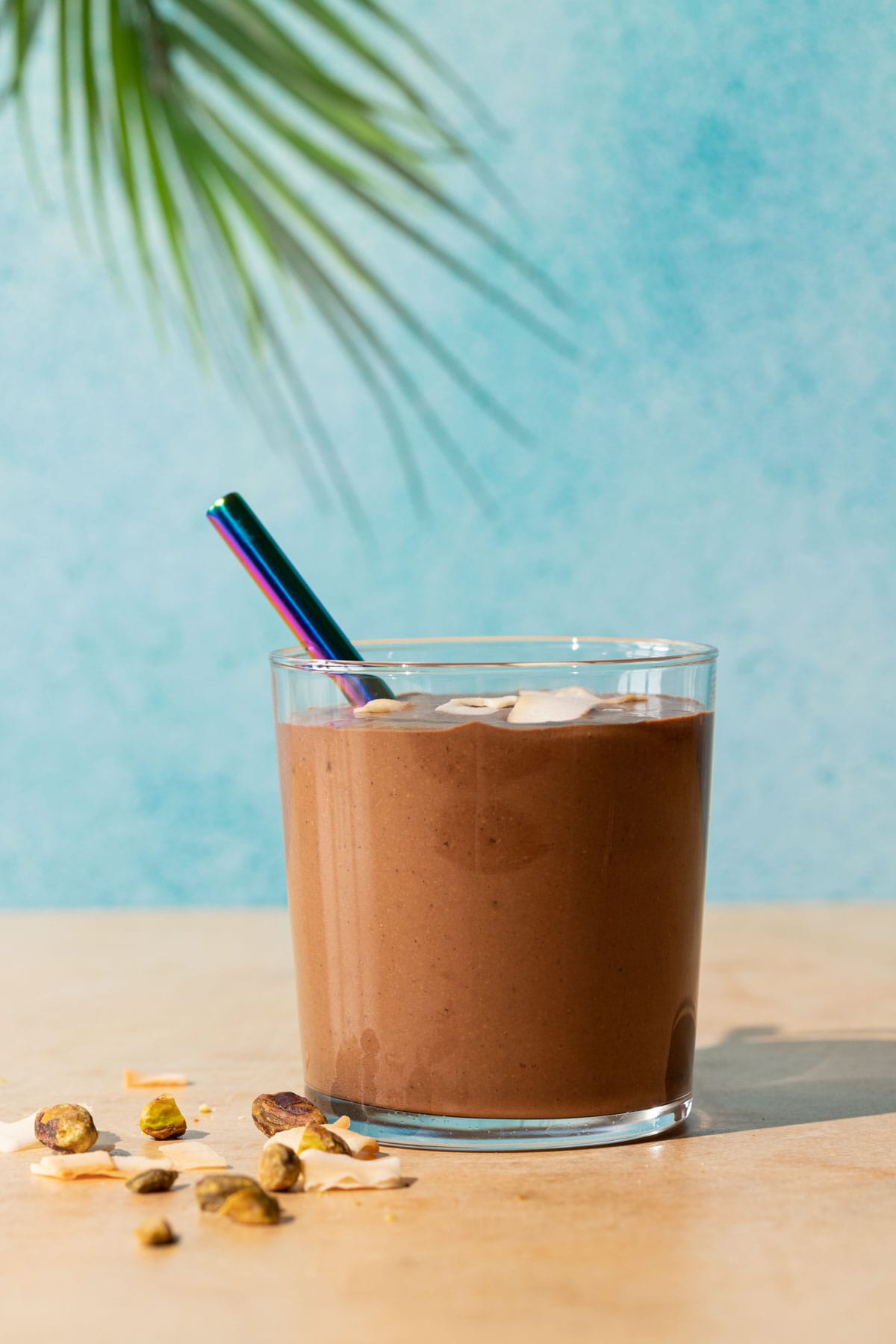 Chocolate Coconut smoothie with toasted coconut chips and pistachios on a yellow and blue background with palm leaf in background