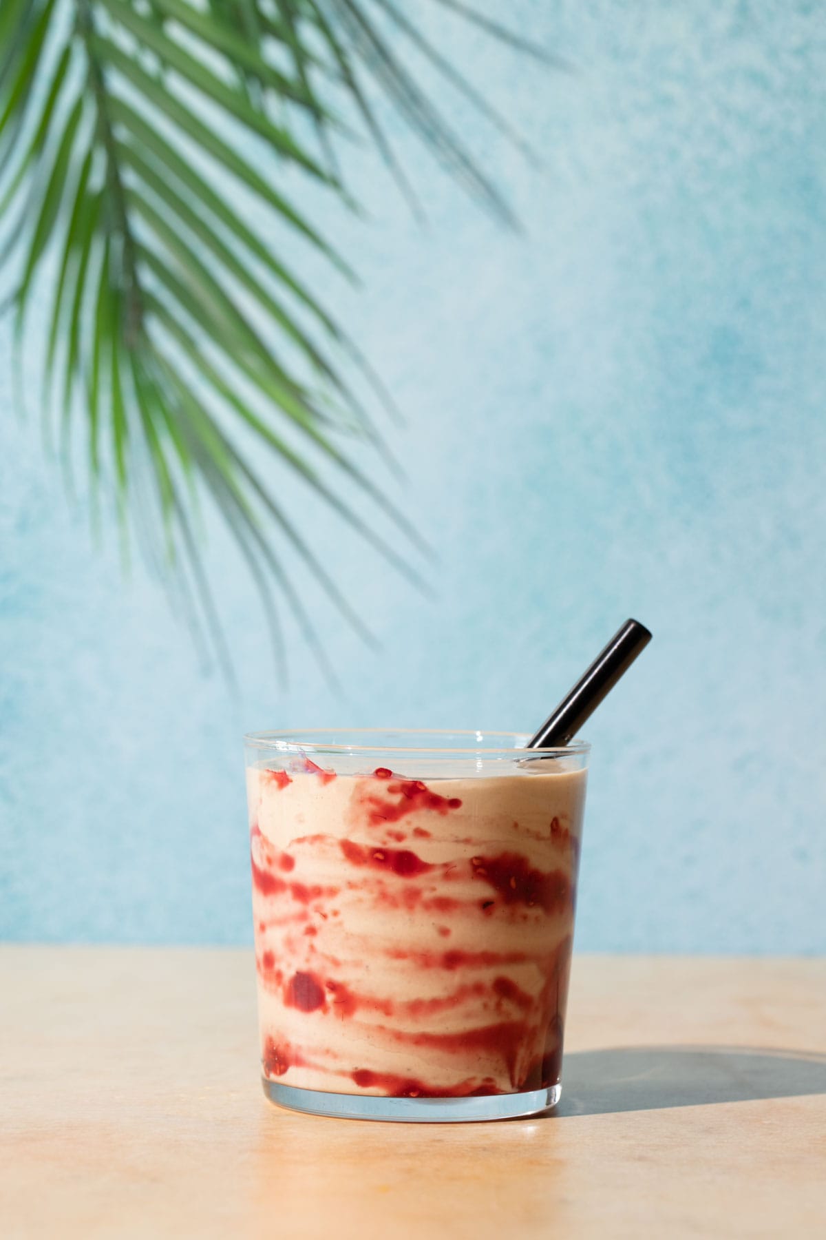 PB&J smoothie with a black straw on a blue and yellow background with a palm leaf overhead
