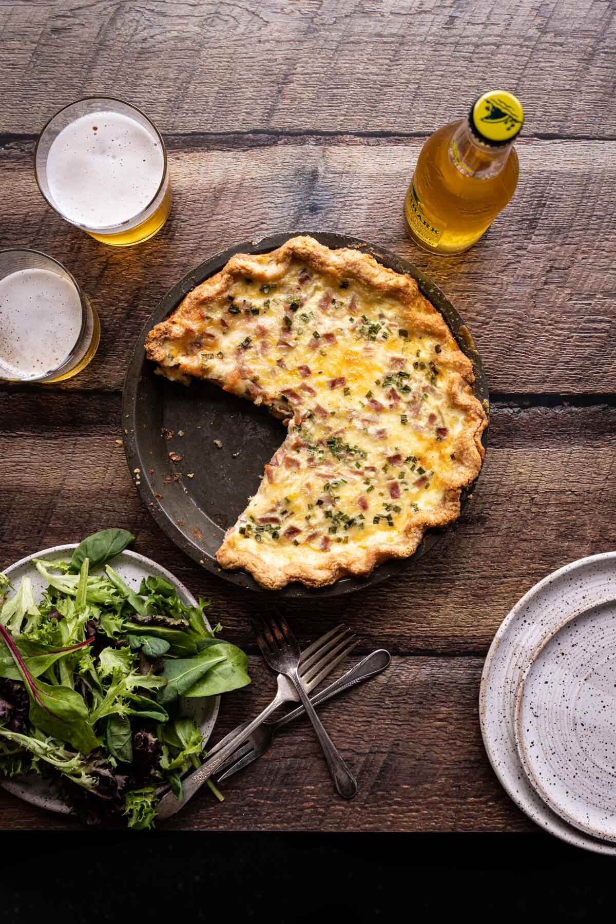 French Beer Quiche with a slice removed sitting beside a salad and two glasses of beer on a wooden table