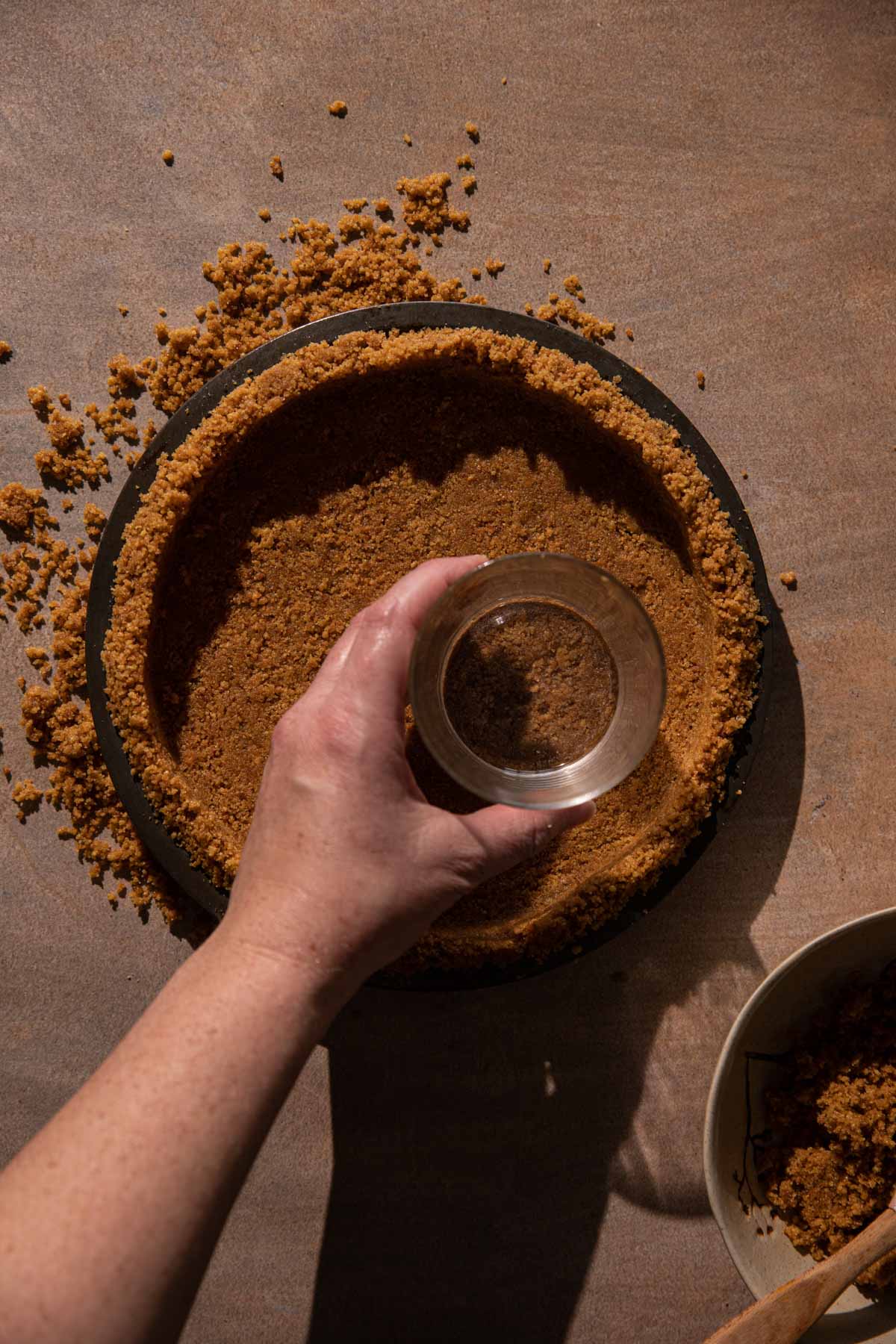 A hand pressing down gingersnap crust in a pie pan with a glass