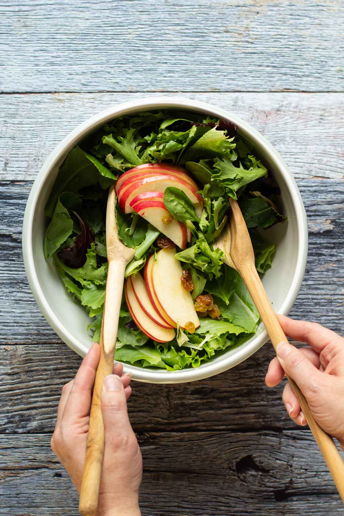 Two hands with wooden spoons mixing apples into a salad in a bowl
