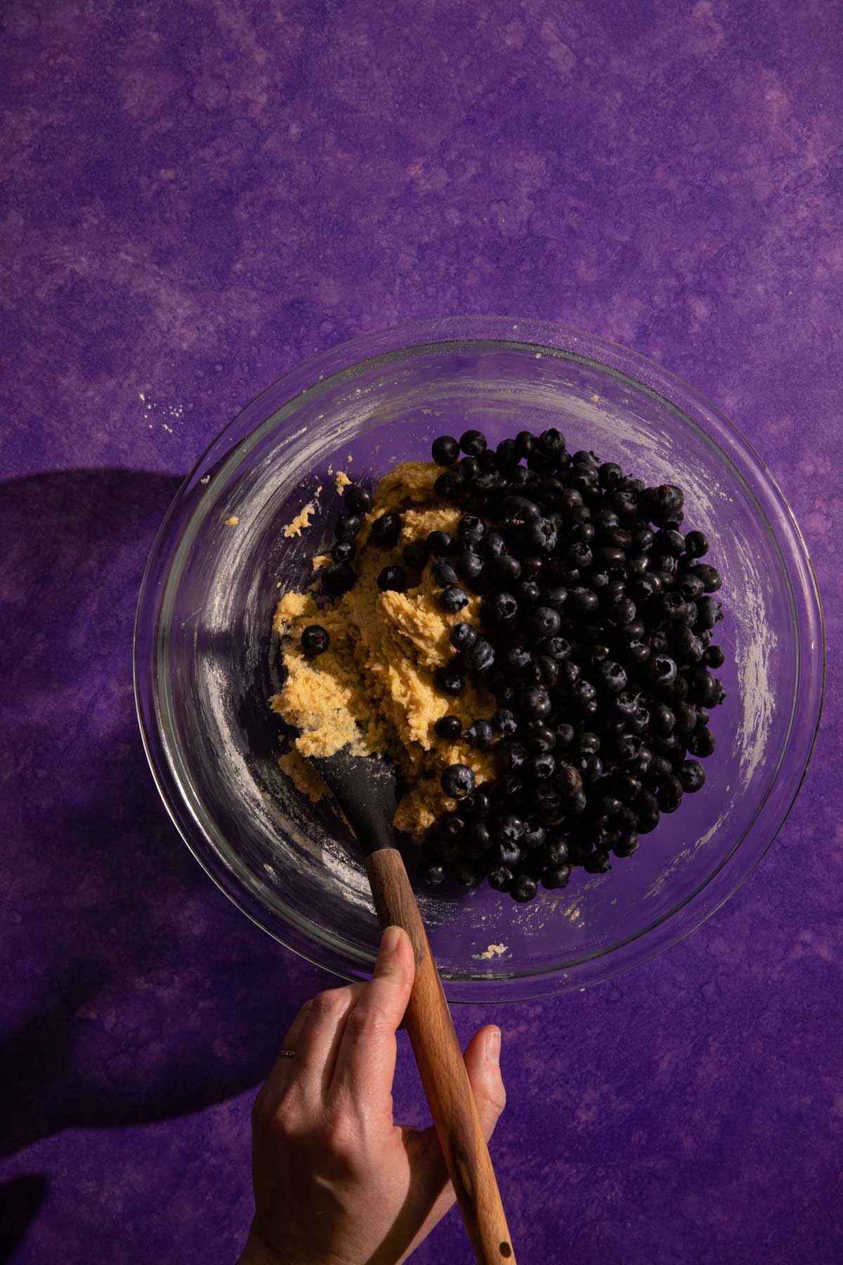 A hand with a spatula stirring blueberry muffin batter 