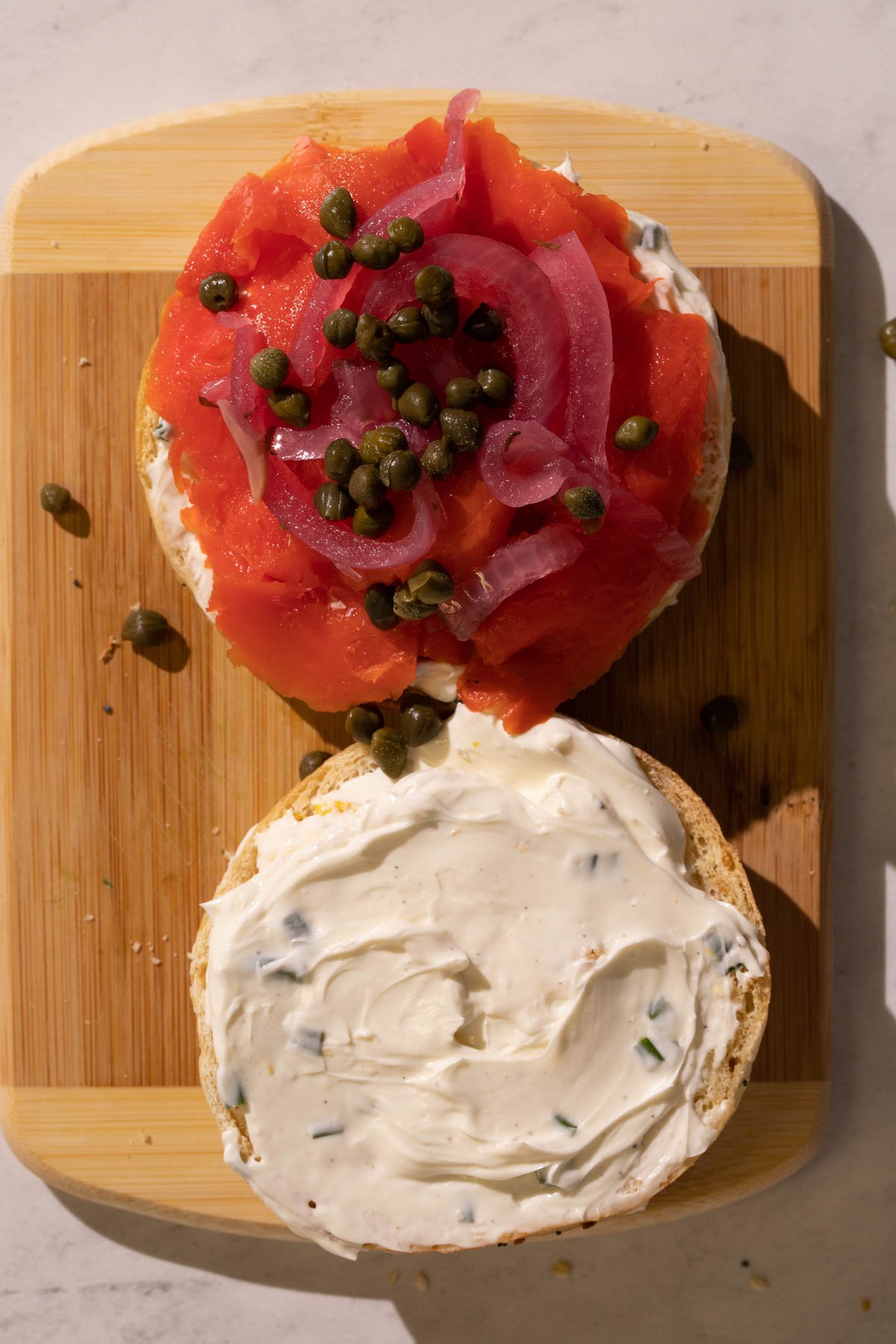 A toasted everything bagel cut in half, covered with cream cheese with chives and fresh lemon, and one half of the bagel, smoked salmon is layered and topped with pickled red onion and capers.