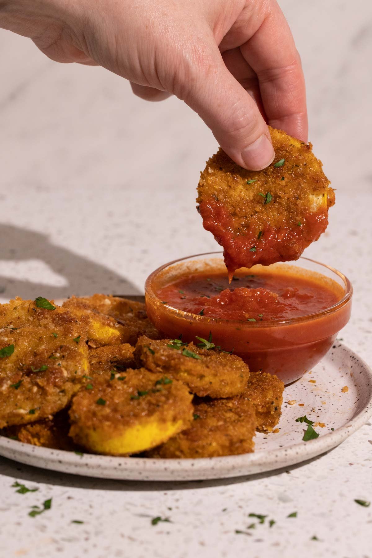 A hand dipping a piece of fried yellow squash in marinara sauce